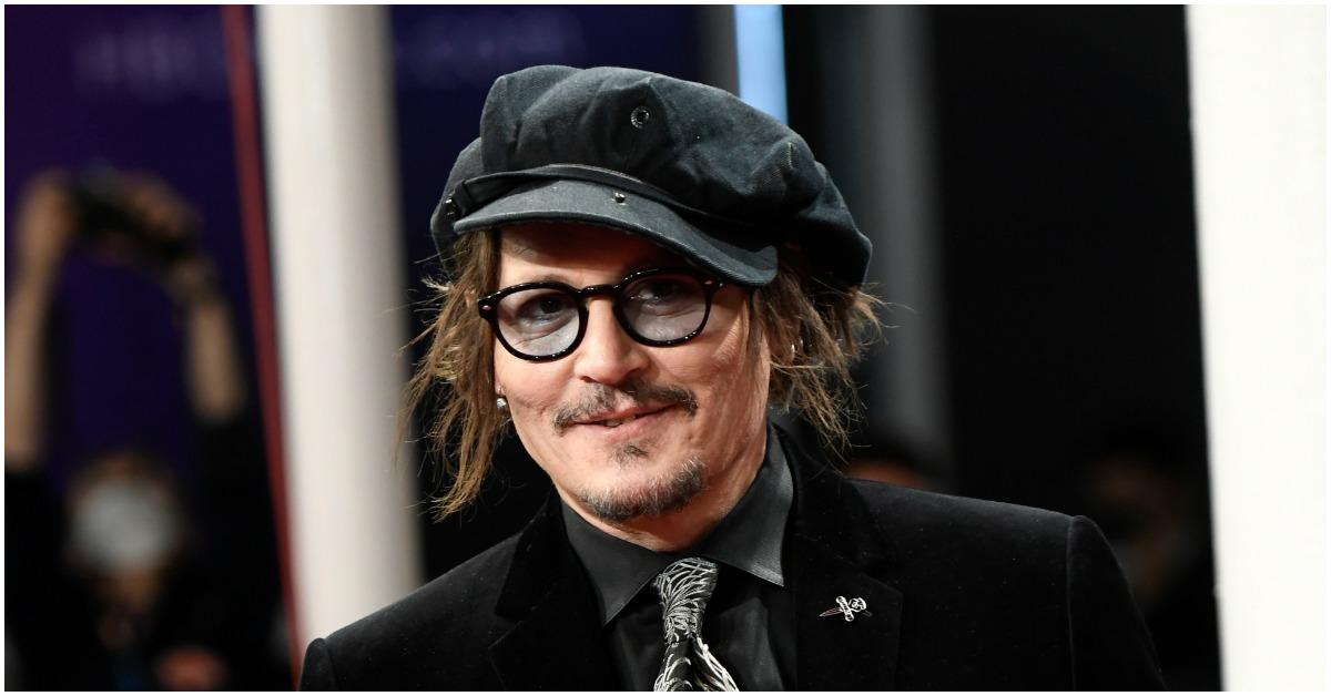 Johnny Depp smiling at a red carpet event wearing a black suit, hat, and glasses.