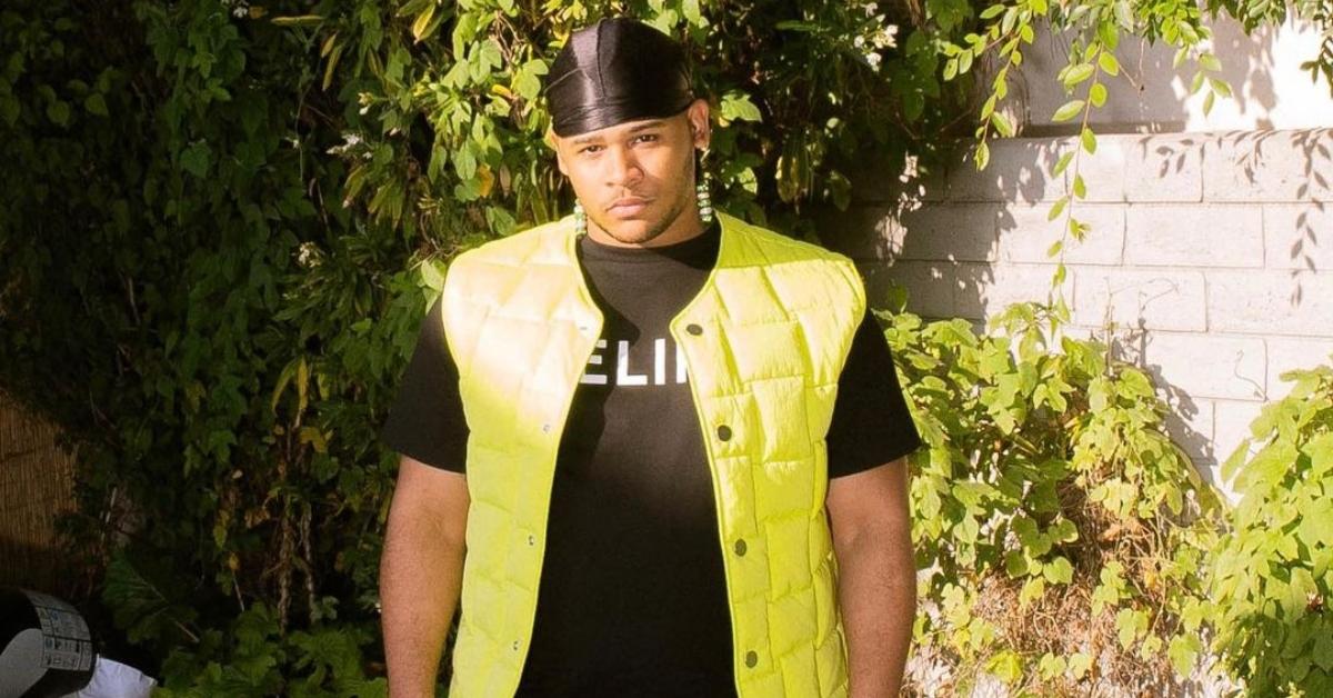 Twitch streamer Fanum wears a neon green vest, black shirt, and black durag, and looks serious next to a stone wall with vines.