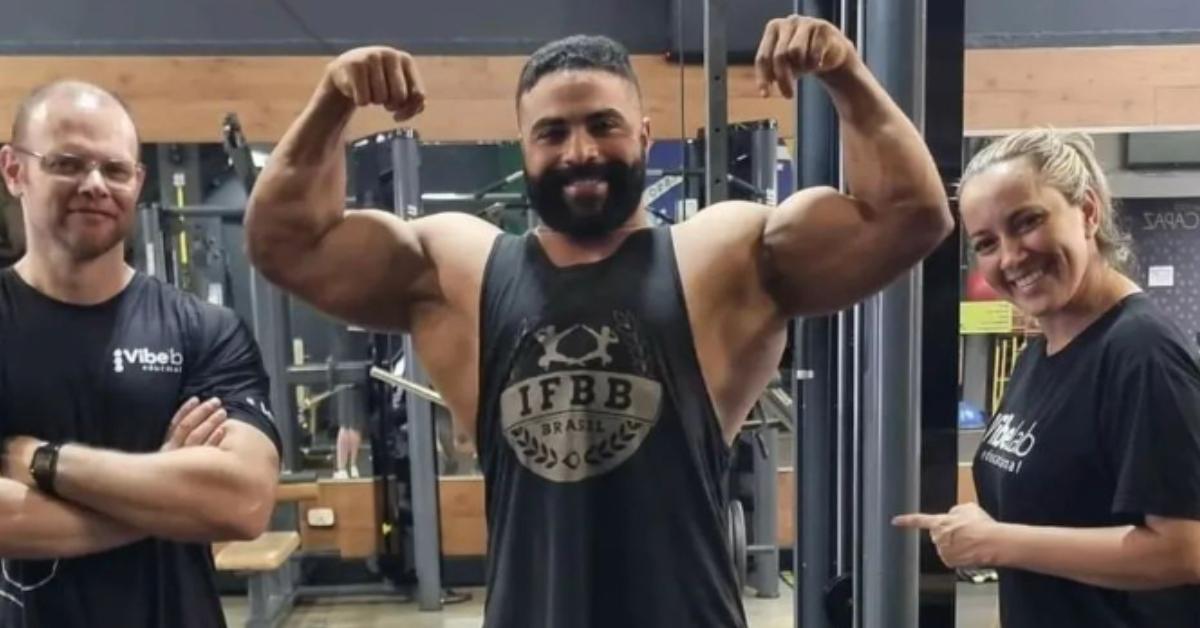 Andre Cavalcanti shows off his muscles in the gym with two other people smiling alongside him.