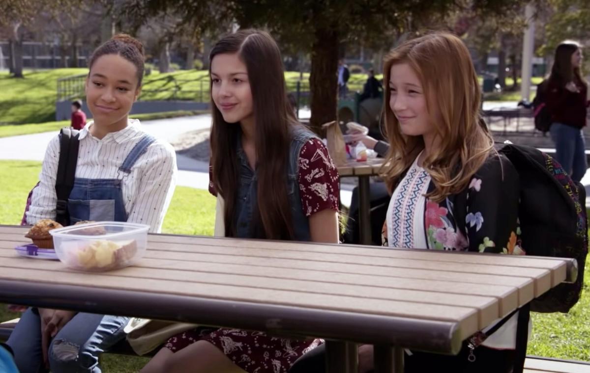 Olivia Rodrigo as a student in 'New Girl,' sitting outside at a picnice table with two other girls