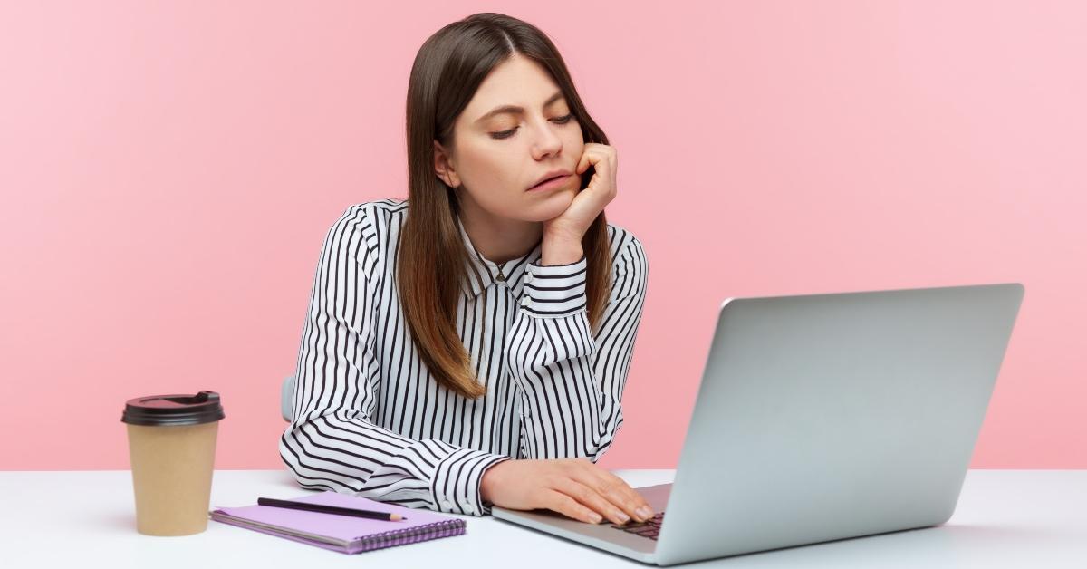 Woman wearing a striped button down shirt looking annoyed sitting at a laptop with coffee, a notepad, and pen.