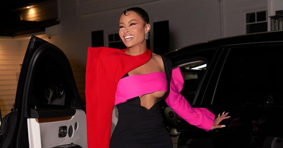 Blac Chyna wears a pink, black and red dress and poses near black SUV