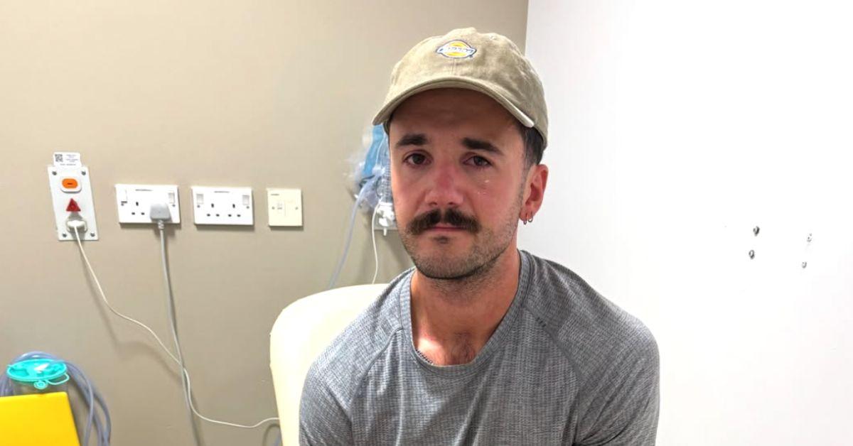 Luke Tarrant in a gray shirt sitting on a hospital bed. 