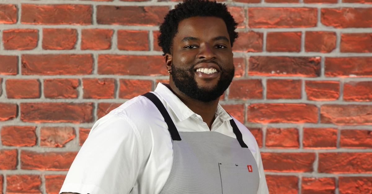 Izahya from Season 3 of 'Next Level Chef' stands in front of a brick wall wearing a gray apron