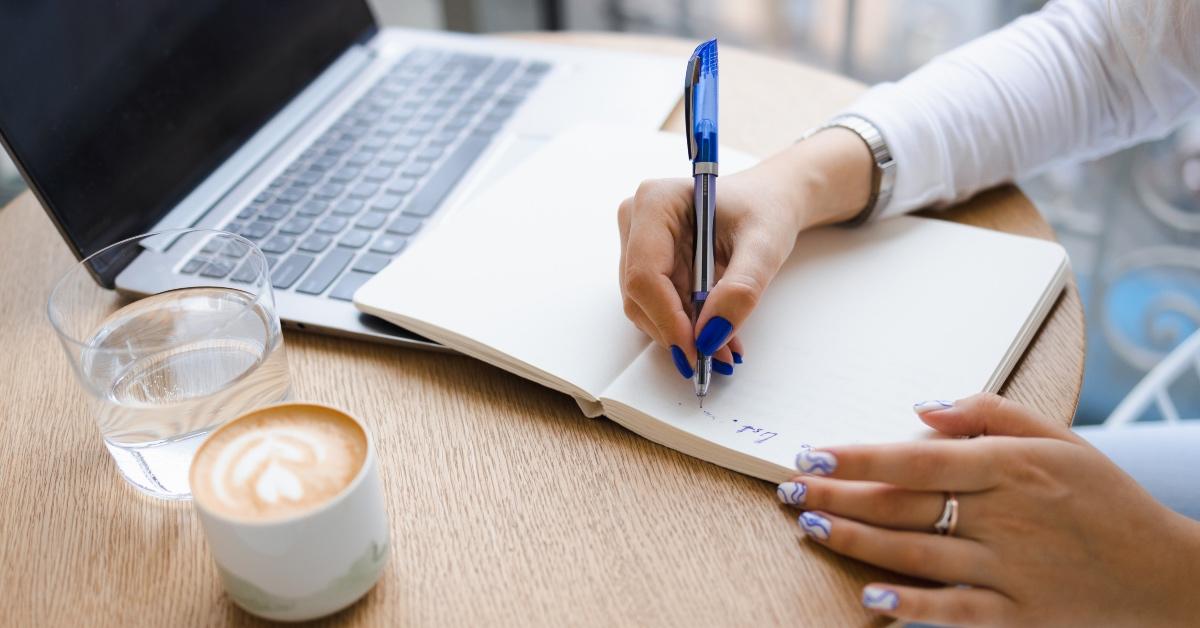 Woman with painted nails writing a book in a notebook while sitting in front of a laptop computer.