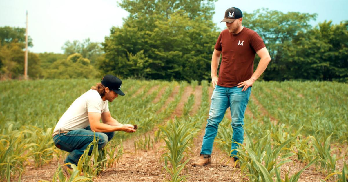 Jesse McBee, clad in a black baseball cap, cream-colored shirt, and jeans, crouches in the field while Cole, sporting a rusty-colored shirt, jeans, and a black baseball cap, looks at him.