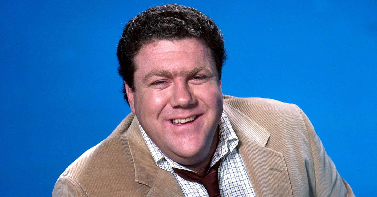 George Wendt smiles and poses in front of a blue background.