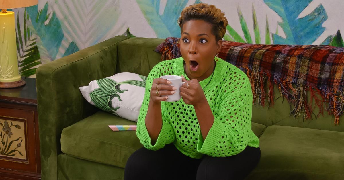Paul/Caress, wearing a neon green sweater and black pants, holds a white mug and has a shocked facial expression while sitting on the couch in Season 6 of 'The Circle.'