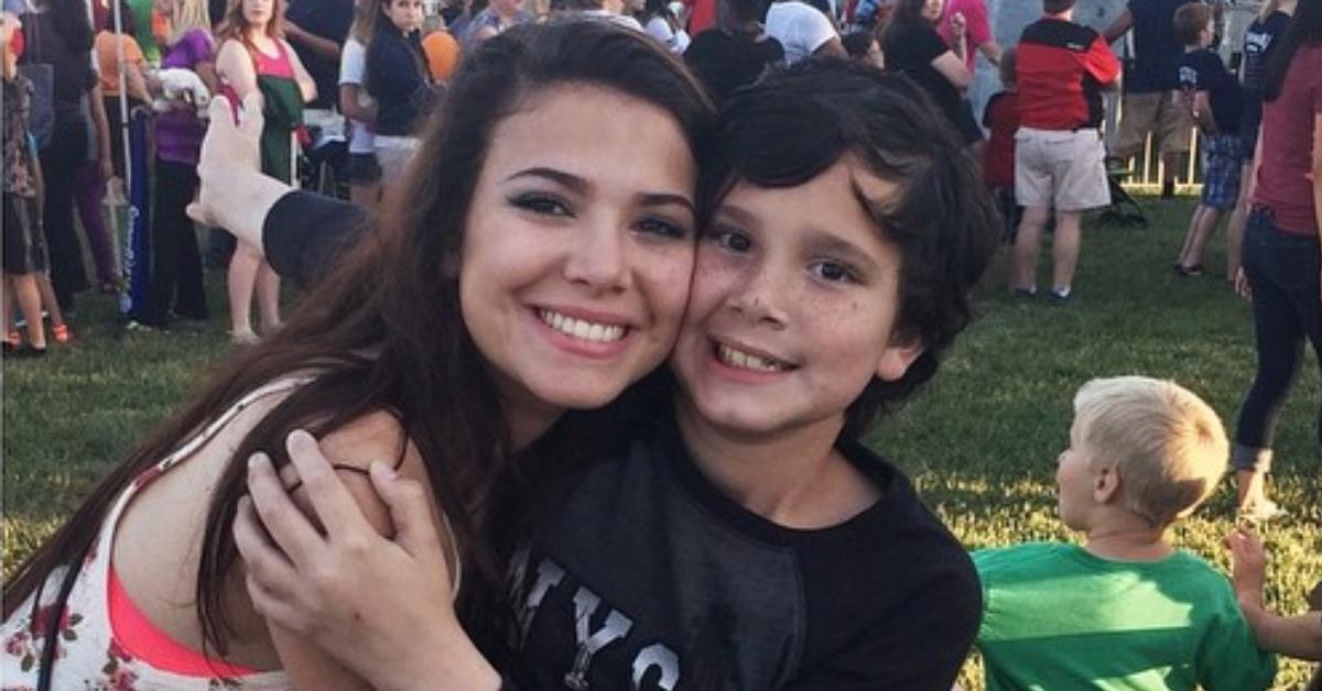 Amy Cortés is wearing a floral tank top as she beams with a smile and embraces her younger brother, David, who is rocking a black long-sleeve graphic tee.