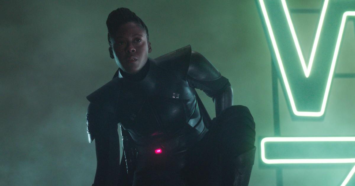 Reva (Moses Ingram) in her black Sith Inquisitor armor, kneels in front of a neon sign. 