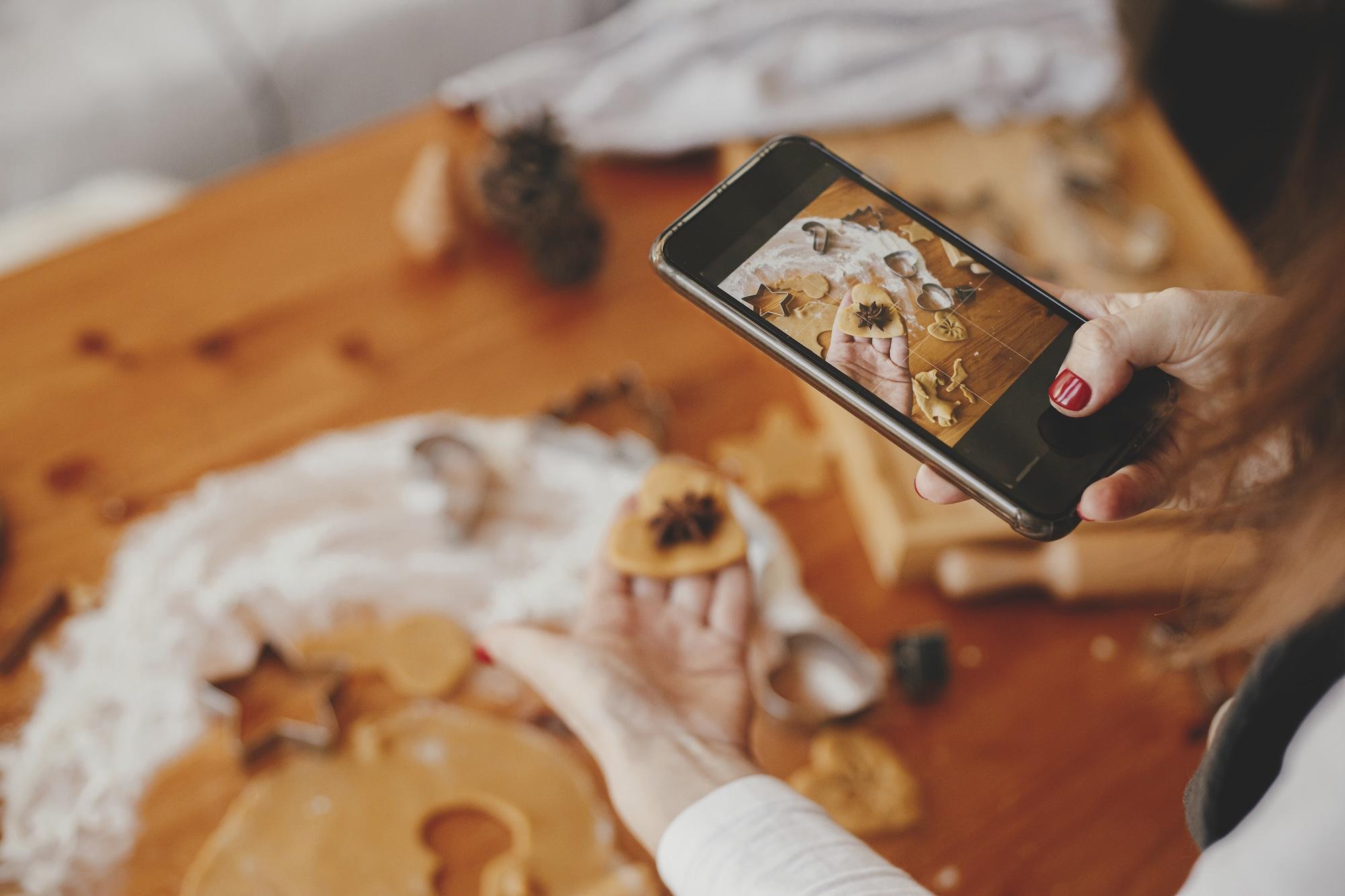 Woman holding phone and taking photo of gingerbread cookies christmas cookies on messy table flat lay. Instagram photo, social media and blog. Holiday preparations 