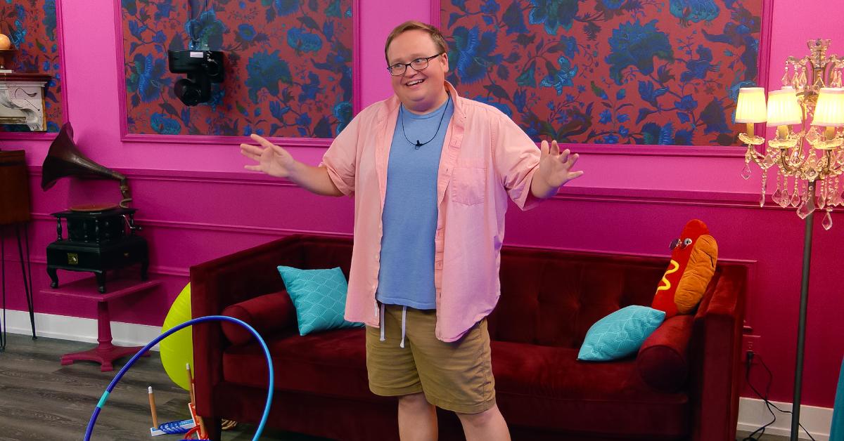 Brandon, in khaki shorts, a blue t-shirt, and pink short-sleeved unbuttoned button-up, stands in front of the red couch in 'The Circle' Season 6.