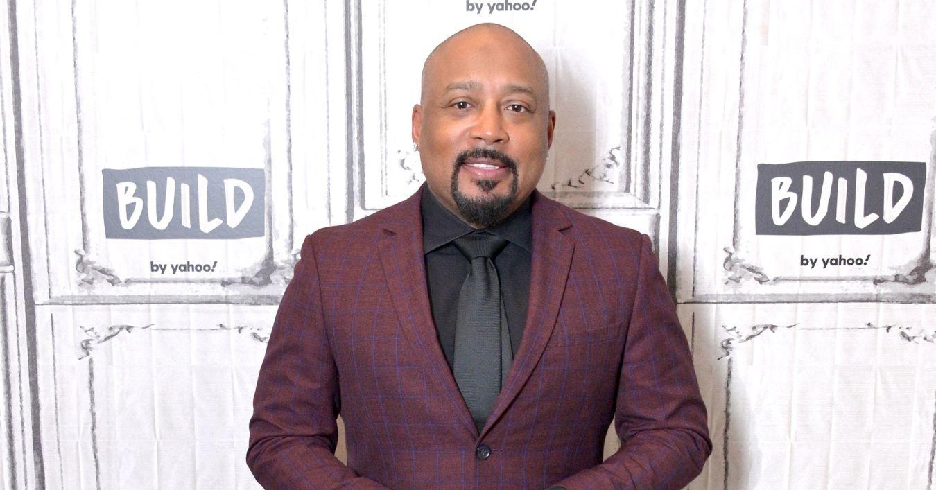Daymond John on the red carpet in a maroon suit with a black tie.