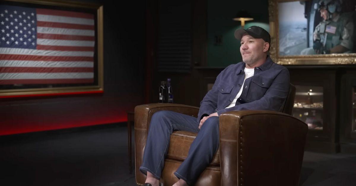 Shawn Ryan sitting in an armchair with an American flag on the wall. 