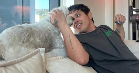 Clinton Kane wears blue nail polish and sits on a couch wearing a gray t-shirt while petting a white dog
