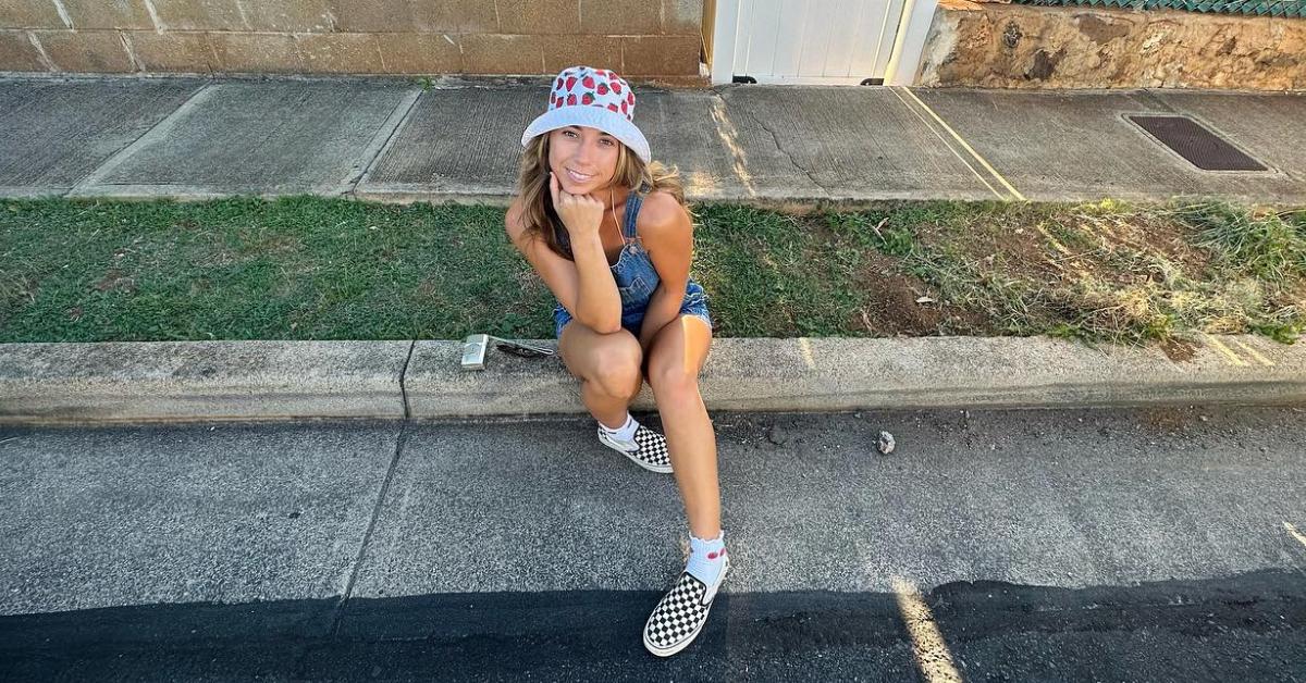 Haleigh Gorman wears a strawberry bucket hat, overalls, and checkerboard-patterned Vans while sitting on a sidewalk.