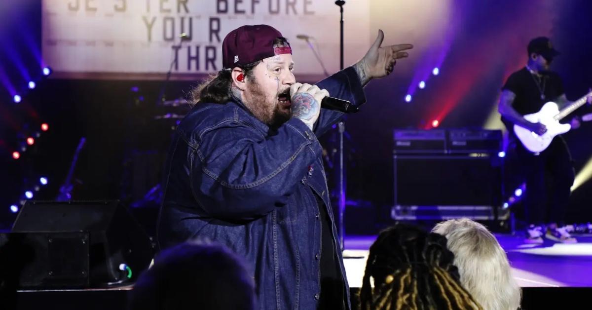 Jelly Roll holds microphone and sings to audience in a jean jacket.