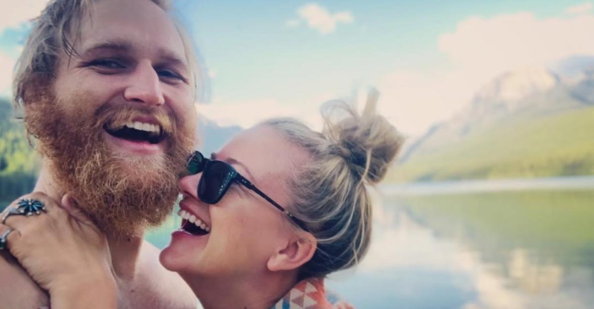 Wyatt Russell and Meredith Hagner laugh and embrace in front of a scenic lake.