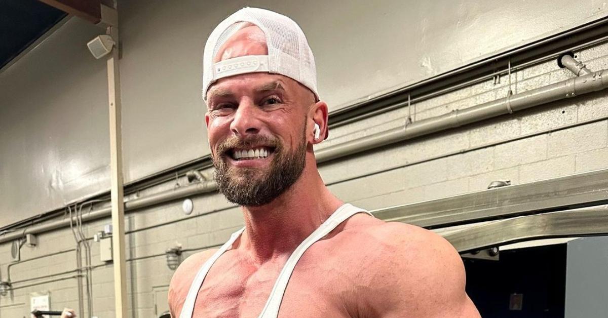 Joey swole wears a white trucker hat backward and a white tank top and smiles at the gym