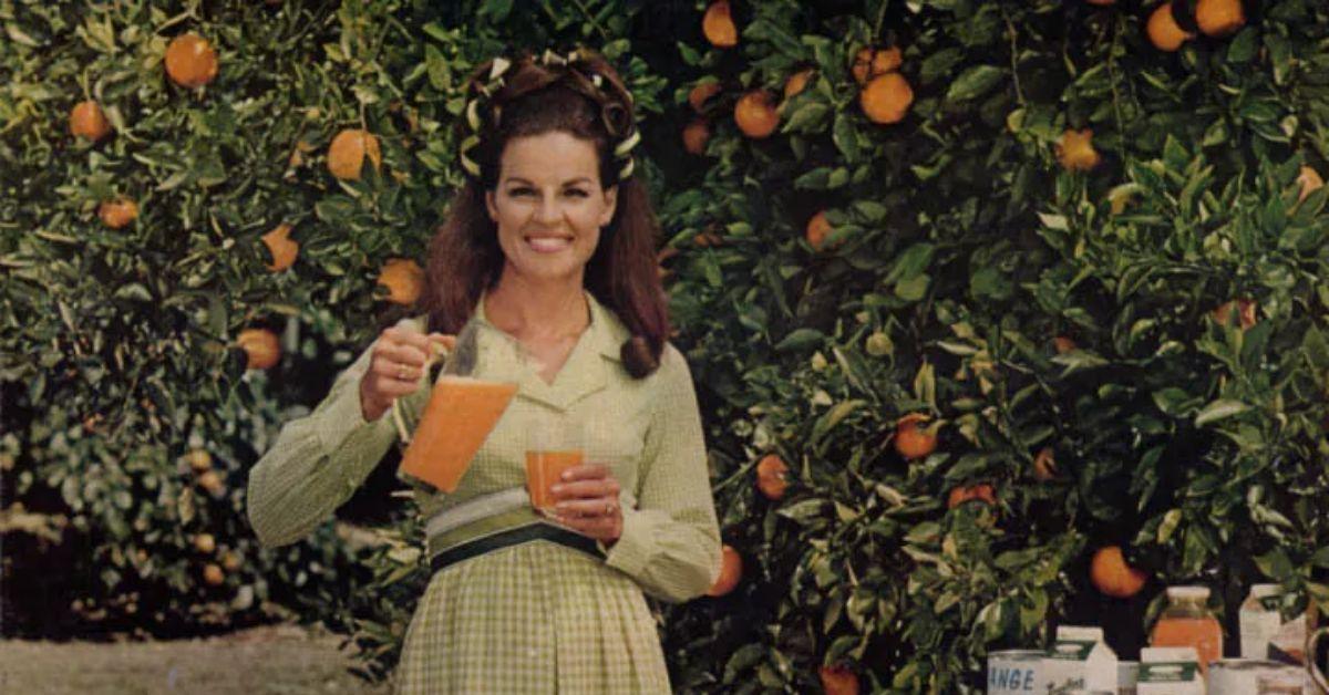 Abita Bryant holding orange juice with oranges behind her. 