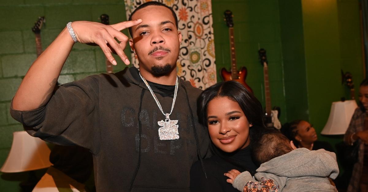 G Herbo wears a black shirt and diamond chain standing next to Taina holding G Herbo's baby. 