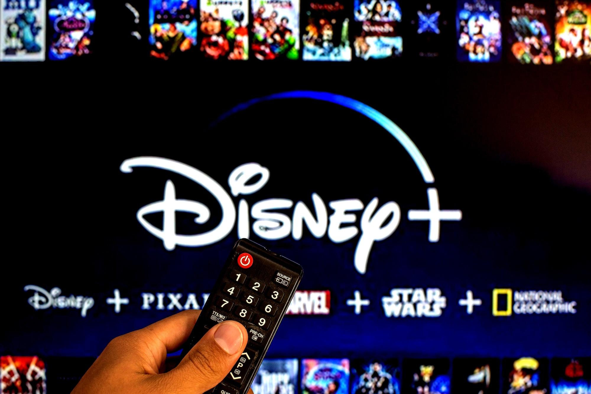 A person holding a TV remote control seen displayed in front of the Disney+ logo