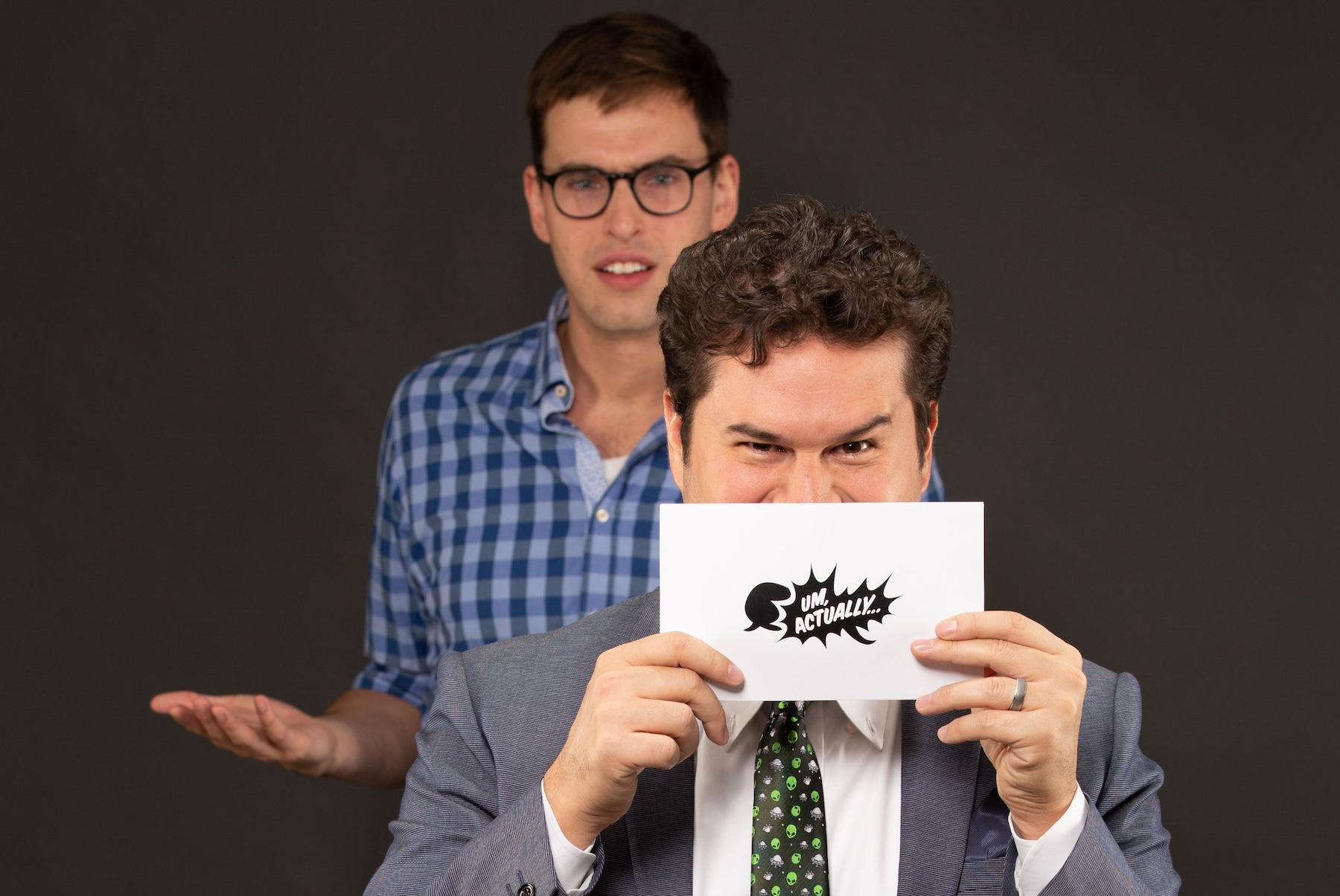 Grant O'Brien looking confused behind Mike Trapp holding up an 'Um, Actually' card in front of his face