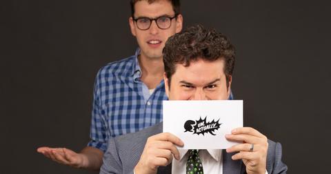 Grant O'Brien looking confused behind Mike Trapp holding up an 'Um, Actually' card in front of his face