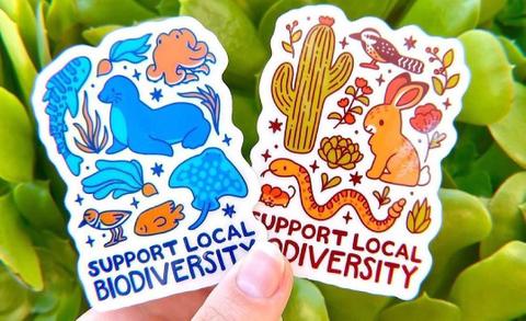 A person holding two stickers that say "support local biodiversity" with various animals.