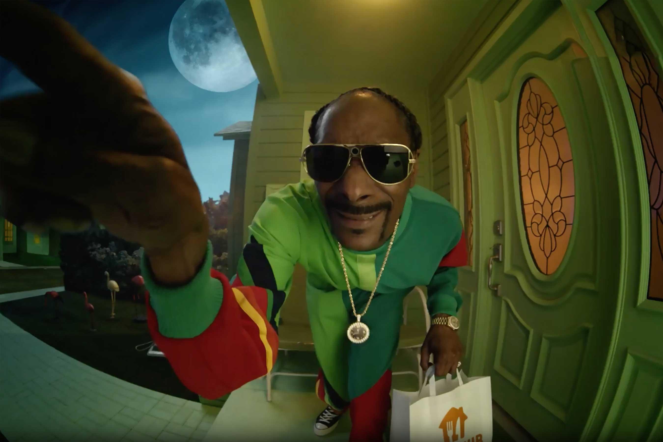 Snoop Dogg wears a green and red track suit and rings the ring doorbell camera holding a white and orange.