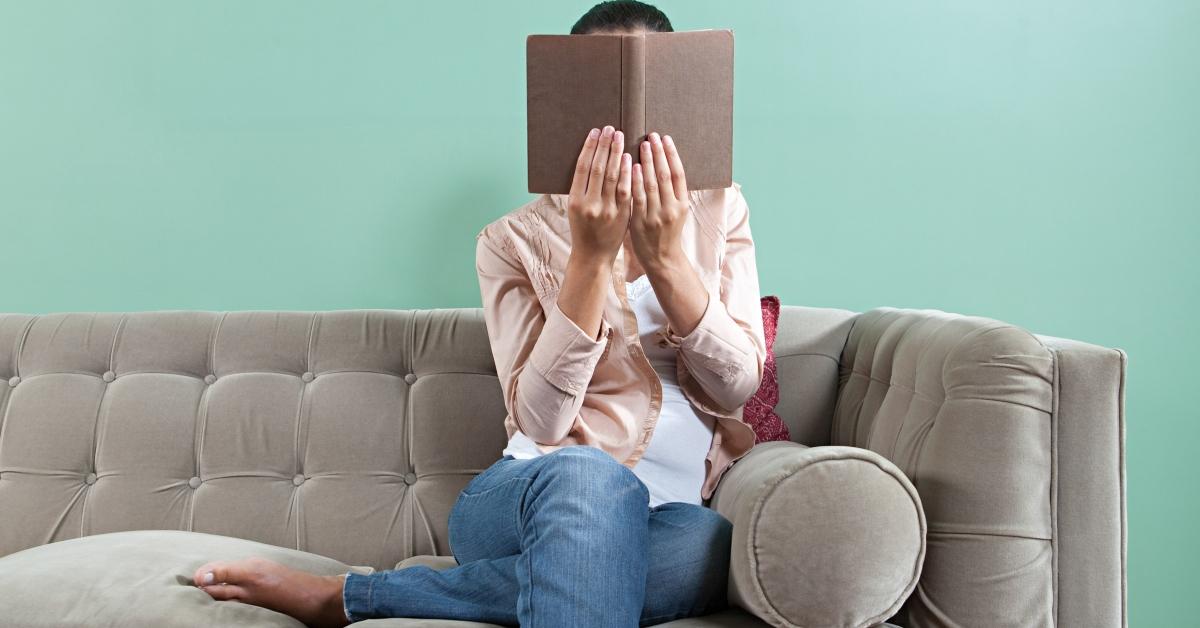 woman holding up book on couch, covering her face with it